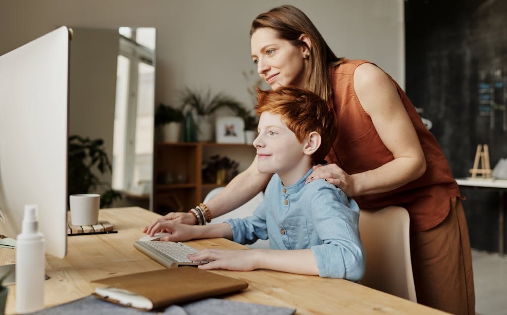 Teaching Kids About Money: Because ‘Mum, Can I Have More?’ Isn’t a Financial Plan 💷😂 2 A mother and her child smiling while using a computer at home, focused on learning.